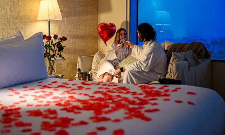 A couple in bathrobes in a romantic room at TOO Hotel, with a panoramic view of Paris, heart-shaped balloons, and rose petals.